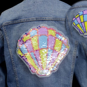 Peut inclure: Une veste en jean avec un grand patch en forme de coquille de mer en paillettes irisées sur le dos. Le patch est rose, bleu et jaune avec un contour blanc.