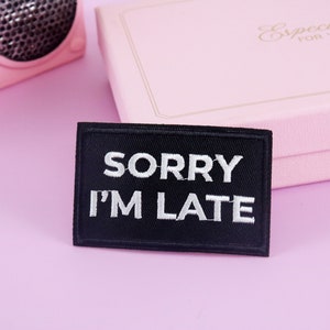 Puede incluir: Un parche negro bordado con texto blanco que dice "SORRY I'M LATE".