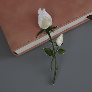 May include: Embroidered white rose patch with green stem and leaves. The patch is placed on a pink notebook with a brown ribbon bookmark. The background is a solid grey surface.