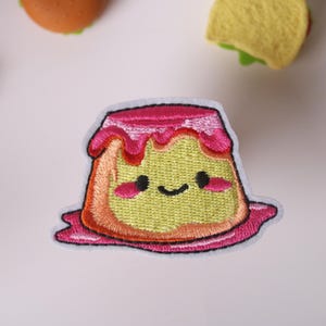 May include: A pink and yellow embroidered patch of a cartoon pudding with a happy face. The pudding has a pink swirl on top.