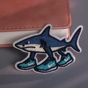 以下が含まれることがあります： 青と白の漫画のサメの刺繍パッチ。白いアクセントのついたターコイズ色のスニーカーを履いています。パッチは白い縁取りがあり、衣類やアクセサリーに適用するように設計されています。サメには足があります。