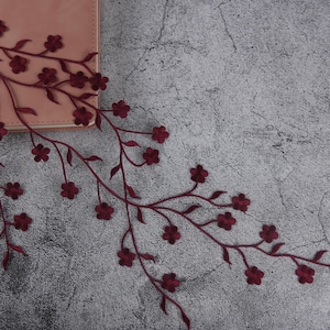 Può includere: Un ricamo floreale bordeaux con piccoli fiori e foglie dettagliate. Il ramo ricamato è posto su una superficie rosa chiaro e uno sfondo grigio strutturato. Questo elemento decorativo potrebbe essere utilizzato per abbellire abiti o lavori artigianali.