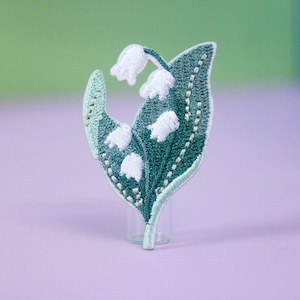 Op de afbeelding: Een wit geborduurde lelietje-van-dalen-bloemenpatch met groene bladeren. De patch bevindt zich op een lichtpaarse achtergrond.