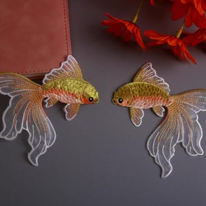 Könnte beinhalten: Zwei gestickte Goldfisch-Aufnäher mit goldenen, orangefarbenen und weißen Details. Die Fische haben detaillierte Flossen und Schwänze und sind vor grauem Hintergrund platziert. Dekorative Aufnäher für Kleidung oder Bastelarbeiten.