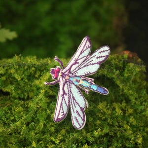 May include: A purple and blue embroidered dragonfly patch with white wings. The patch is on a green mossy surface.
