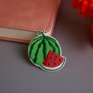 May include: Embroidered watermelon patch featuring a green rind and a red, seeded slice. The patch is white-bordered and sits on a gray surface, next to a closed pink notebook and orange flowers.