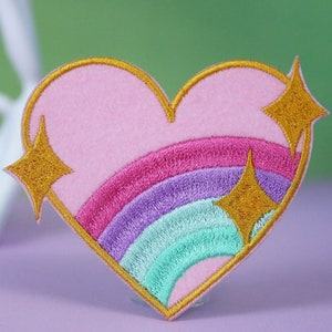 Könnte beinhalten: Rosa Herzform-Patch aus Filz mit einem Regenbogen und goldenen Glitzersternen, die darauf gestickt sind.