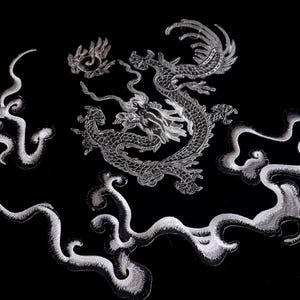 Pode incluir: Um desenho de dragão bordado em prata sobre um fundo preto. O dragão é rodeado por nuvens em espiral.