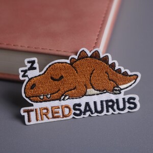 Op de afbeelding: Geborduurde patch met een bruine dinosaurus die slaapt met gesloten ogen en het woord "TIREDSAURUS" eronder. De patch heeft een witte rand en ligt op een grijze ondergrond.