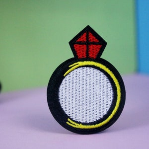 Puede incluir: Un parche bordado en forma de anillo con un diseño en blanco y negro y un diamante rojo en la parte superior. El anillo está rodeado de un contorno amarillo.