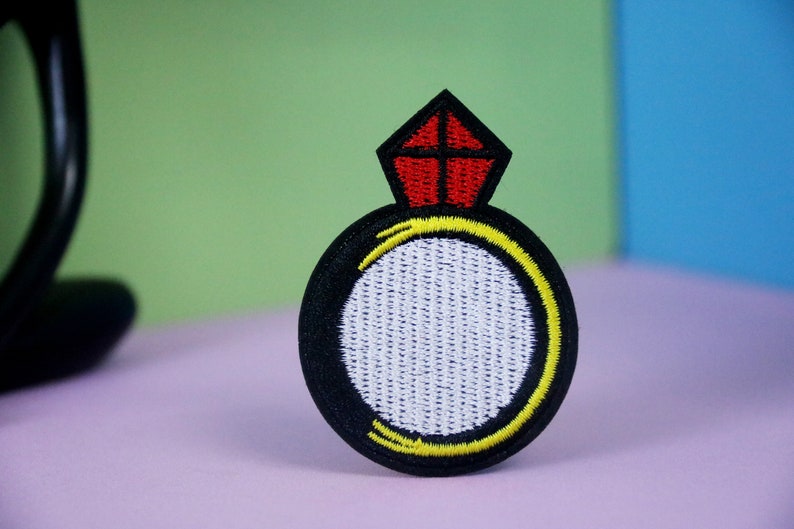 May include: A black and white embroidered ring with a red diamond on top. The ring is surrounded by a yellow outline.