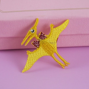 May include: A yellow embroidered iron-on patch of a cartoon pterodactyl with red accents.