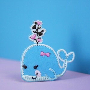 Könnte beinhalten: Ein gestickter blauer Wal-Aufnäher mit einer rosa Schleife und einer rosa Blume oben. Der Wal hat weiße Nähte an den Rändern und schwarze Augen.