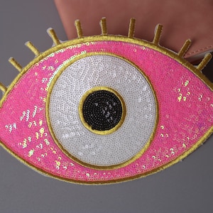 Puede incluir: Un parche en forma de ojo rosa y blanco con ribete dorado y pupila negra. El parche está hecho de lentejuelas y es perfecto para agregar un toque de fantasía a la ropa o los accesorios.