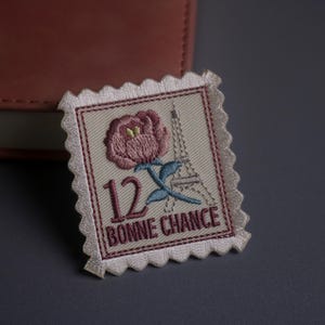 Peut inclure: Écusson brodé en forme de timbre-poste. Le motif représente une rose rose, la Tour Eiffel, le chiffre 12 et les mots "BONNE CHANCE" en bordeaux. L'écusson a un bord festonné et est posé sur un fond neutre.
