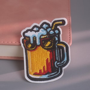 Könnte beinhalten: Ein gestickter Aufnäher in Form eines gelben Bierkrugs mit dickem weißem Schaum, schwarzem Henkel und schwarzem Strohhalm. Der Krug trägt eine schwarze Sonnenbrille mit gelben Gläsern und ist mit weißem Garn auf dunklem Grund umrandet.