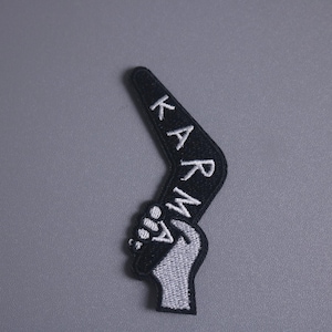 Könnte beinhalten: Schwarzer, gestickter Aufnäher mit dem Wort "KARMA" in weißer Schrift. Der Aufnäher hat die Form eines Bumerangs mit einer geballten Faust am unteren Ende. Der Aufnäher befindet sich auf grauem Hintergrund.