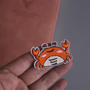 Op de afbeelding: Geborduurde patch met een cartoonkrab met een oranje lichaam, witte accenten en een lachend gezicht. De krab heeft grote ogen en oranje klauwen. Geschikt voor kleding of accessoires.