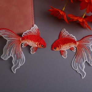 Op de afbeelding: Twee geborduurde goudvispatches met rode en witte details. De vissen hebben vloeiende staartvinnen en staan tegen een grijze achtergrond. Decoratieve bloemelementen zijn zichtbaar.