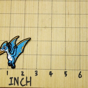 Pode incluir: Um remendo bordado com um pterod&aacute;ctilo de desenho animado em azul e amarelo. O remendo est&aacute; sobre uma superf&iacute;cie de madeira com um padr&atilde;o de grade e marcas em polegadas.