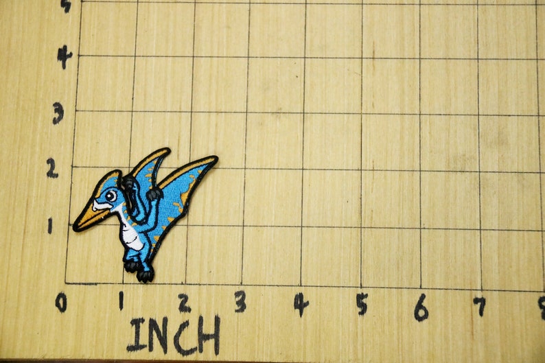 Pode incluir: Um remendo bordado com um pterod&aacute;ctilo de desenho animado em azul e amarelo. O remendo est&aacute; sobre uma superf&iacute;cie de madeira com um padr&atilde;o de grade e marcas em polegadas.