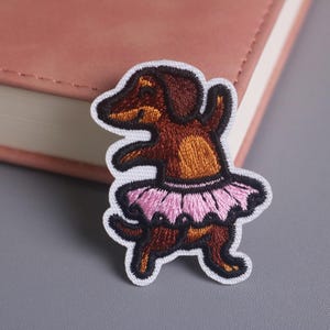 May include: Embroidered patch of a dachshund dog in a ballet pose, wearing a pink tutu. The patch has a white border and is set against a pink book and grey surface. The dog is brown and tan.
