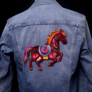 Puede incluir: Chaqueta vaquera con un diseño de caballo bordado colorido en la espalda. El caballo presenta detalles rojos, naranjas, rosas y azules. La chaqueta es de un azul medio.