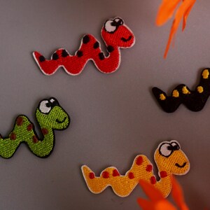 May include: Four embroidered snake patches in red, green, yellow, and black. Each patch has a white outline and a smiling face. The red snake has black spots, the green has red spots, the yellow has orange spots, and the black has yellow spots. These patches are suitable for clothing or crafts.