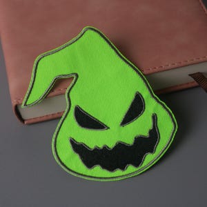 Könnte beinhalten: Ein grüner Filz-Patch mit einem schwarzen gestickten Gesicht eines Charakters mit einem spitzen Hut und einem breiten, zahnigen Grinsen.
