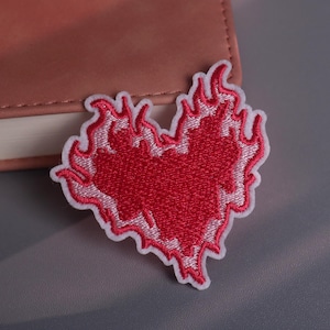 May include: Embroidered patch in the shape of a heart engulfed in flames. The heart is red with white outlining, and the flames are also white. The patch is placed on a gray surface next to a pink notebook.