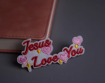 Op de afbeelding: Geborduurde patch met de woorden "Jesus Love You" in rood schrift. De patch heeft roze harten en gele sterren op een witte achtergrond. Op de achtergrond staat een roze leren notitieboek.