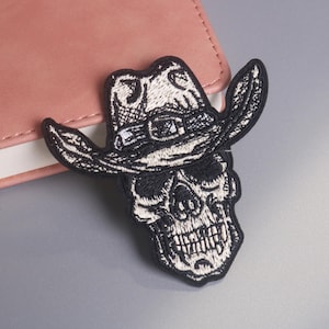 May include: Embroidered patch featuring a skull wearing a cowboy hat. The patch is black and white, with detailed stitching. The skull has visible teeth and eye sockets. The hat has a buckle and brim. The patch is likely for clothing or accessories.