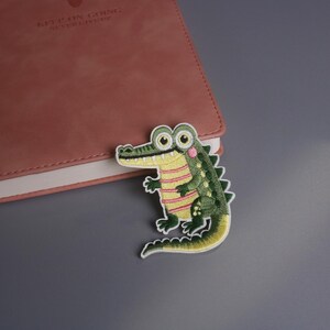 May include: An embroidered alligator patch with a green body, yellow striped shirt, and pink accents. The patch is placed next to a closed, pink leather notebook on a gray surface. The alligator has large, round eyes and a smiling expression.