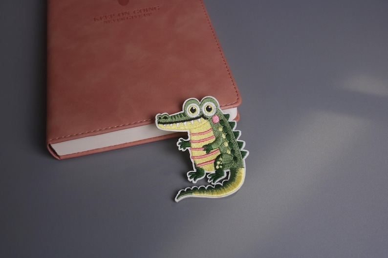 May include: An embroidered alligator patch with a green body, yellow striped shirt, and pink accents. The patch is placed next to a closed, pink leather notebook on a gray surface. The alligator has large, round eyes and a smiling expression.