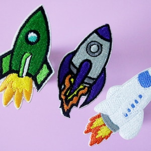 May include: Three embroidered iron-on patches of rockets in different colours. The first rocket is green with a yellow flame. The second rocket is grey with a purple body and an orange flame. The third rocket is white with a blue nose cone and an orange flame.