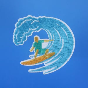 Może przedstawiać: Naszywka haftowana przedstawiająca surfera jadącego na fali. Fala jest niebieska i biała, a surfer ma na sobie zieloną koszulę i niebieskie spodenki. Naszywka idealnie nadaje się do dodania odrobiny plażowego stylu do ubrań lub akcesori&oacute;w.