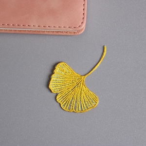 May include: A yellow embroidered ginkgo leaf patch on a gray surface. The patch is detailed with fine stitching, showcasing the leaf's veins. A pink leather item is partially visible in the upper left corner.