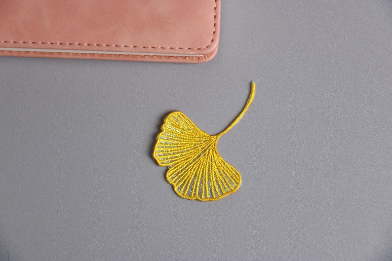 May include: A yellow embroidered ginkgo leaf patch on a gray surface. The patch is detailed with fine stitching, showcasing the leaf's veins. A pink leather item is partially visible in the upper left corner.