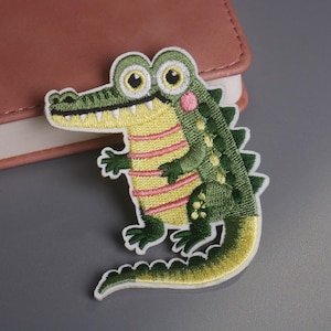 May include: Embroidered alligator patch with a green body, yellow belly with pink stripes, and a white border. The alligator has large eyes, a pink cheek, and a detailed design. This patch is suitable for clothing or accessories.