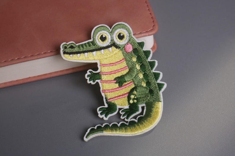 May include: Embroidered alligator patch with a green body, yellow belly with pink stripes, and a white border. The alligator has large eyes, a pink cheek, and a detailed design. This patch is suitable for clothing or accessories.
