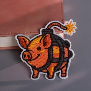 Op de afbeelding: Geborduurde patch met een oranje varken met een zwarte bom. Het varken heeft een lachend gezicht en is wit omlijnd. De bom heeft een aangestoken lont met een gele explosie. De patch ligt op een grijze ondergrond.