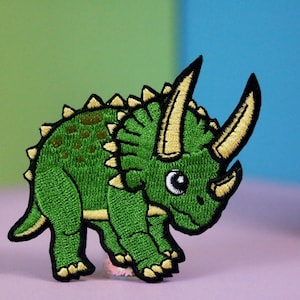 May include: Green embroidered iron-on patch of a cartoon triceratops dinosaur with yellow horns and a black outline.