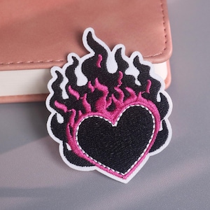 May include: Embroidered patch featuring a black heart with a pink outline and flames. The flames are black with pink accents, and the patch has a white border. The patch is likely for clothing or accessories.