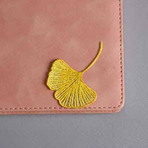 May include: A dusty rose-colored leather notebook with a yellow embroidered ginkgo leaf design in the upper corner. The notebook has a stitched border and is set against a gray background.