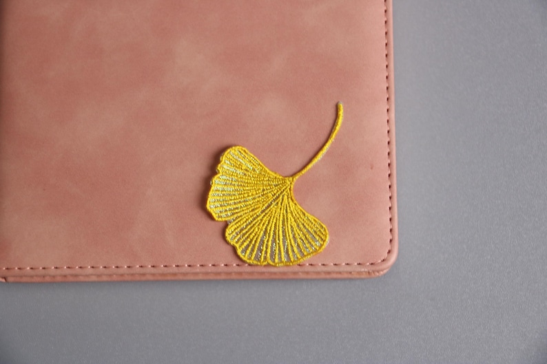 May include: A dusty rose-colored leather notebook with a yellow embroidered ginkgo leaf design in the upper corner. The notebook has a stitched border and is set against a gray background.