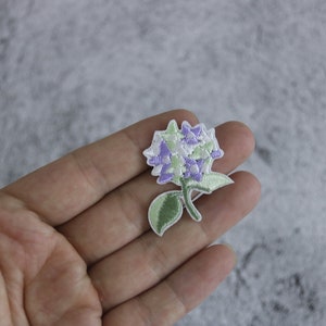 Op de afbeelding: Een geborduurde bloemenpatch in wit en paars met groene bladeren.