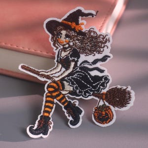 May include: An embroidered patch featuring a witch with brown hair wearing a black hat with an orange ribbon, a black dress, and orange and black striped stockings. The witch rides a brown broomstick and carries a small orange jack-o'-lantern.