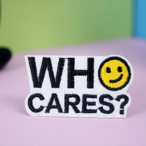 Può includere: Toppa in feltro bianco con testo ricamato in nero che recita "WHO CARES?" e una faccina gialla sorridente.