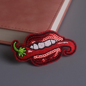 May include: A red sequined patch in the shape of a mouth with a chili pepper. The mouth has white teeth and is outlined in black. The chili pepper has a green stem. The patch is on a gray surface.
