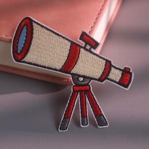 May include: An embroidered patch featuring a beige telescope with red accents and a blue lens, mounted on a red tripod stand with grey feet. The design is outlined in black thread, creating a clear and defined shape for this astronomical instrument.